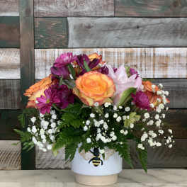 Compact arrangement of orange roses and purple flowers with baby’s breath in a white bee vase.