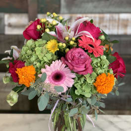 Mixed bouquet of pink roses, gerbera daisies, lilies, marigolds, and green hydrangeas in a clear glass vase