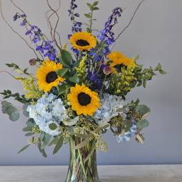 Tall arrangement of yellow sunflowers, blue hydrangeas, and delphinium in a clear glass vase