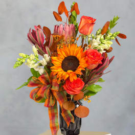 Orange sunflower and roses in a black vase with a plaid ribbon