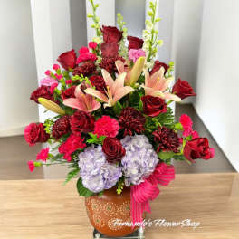 Large mixed arrangement of red roses, pink lilies, bright carnations, and lavender hydrangeas in an orange pot