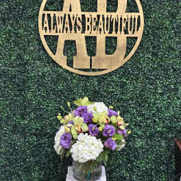 Purple and white floral arrangement in a glass vase beneath a gold monogram sign