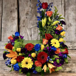 Tall and low arrangement of bright roses, lilies, gerbera daisies, and other blooms in a rectangular container.
