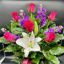 Arrangement of hot pink roses, purple stock, and white lilies in a purple vase with a pink bow