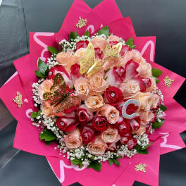 Round bouquet of red and peach roses wrapped in bright pink paper with gold butterfly accents.