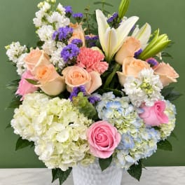 Mixed bouquet of roses, lilies, hydrangeas, and purple filler flowers in a white vase