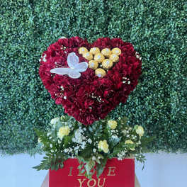 Heart-shaped red carnation arrangement with chocolates and a butterfly topper