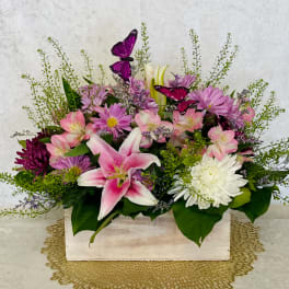Pink lilies and purple daisies arranged in a white wooden box with butterfly picks.