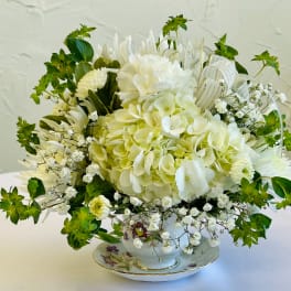 White floral arrangement in a teacup-style vase with green accents and ribbon