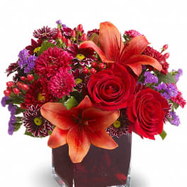 Compact arrangement of red roses, orange lilies, and mixed blooms in a red glass cube vase.