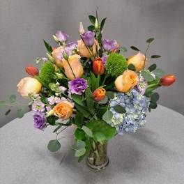 Bouquet of peach roses, purple lisianthus, and blue hydrangea in a glass vase