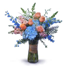 Tall vase arrangement with blue hydrangeas, peach roses, and blue delphinium in a clear glass vase.