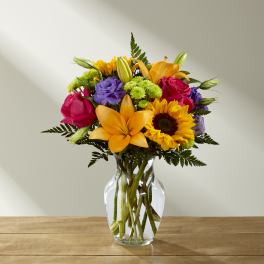 Mixed bouquet of orange lilies, pink roses, purple blooms, and a sunflower in a clear glass vase.