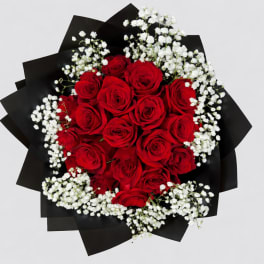 Round bouquet of red roses and white filler flowers wrapped in black paper.