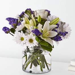 White lilies and daisies with purple filler in a clear glass vase