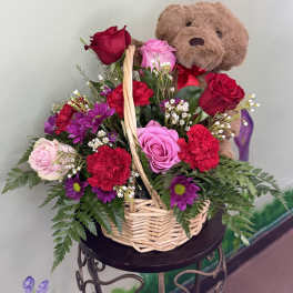 Basket arrangement of red and pink roses, red carnations, purple daisies, and a plush teddy bear.