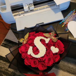 Round bouquet of red roses with a white floral letter S and gold butterflies in black wrap.