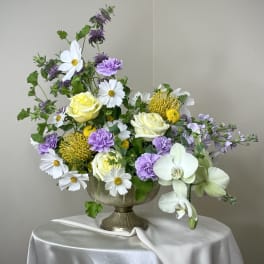 Garden-style arrangement of white, yellow, and lavender flowers in a metal pedestal bowl on a draped table.