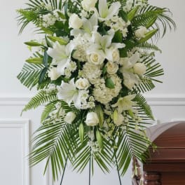 Tall white standing spray of lilies and roses on an easel beside a wooden casket.