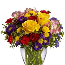 Bright mixed bouquet of yellow roses, red carnations, and purple and yellow blossoms in a clear glass vase