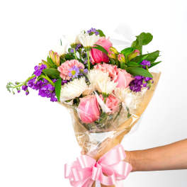 Handheld bouquet of pink, white, and purple mixed flowers wrapped in cellophane with a large pink bow