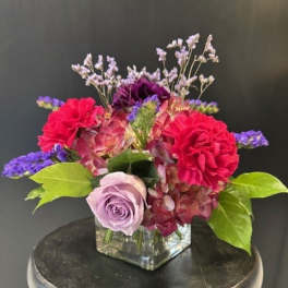 Low arrangement of pink and purple flowers with a lavender rose in a square glass vase.