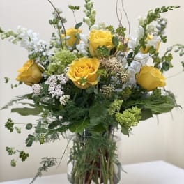 Tall arrangement of yellow roses, white snapdragons, and green accents in a clear glass vase