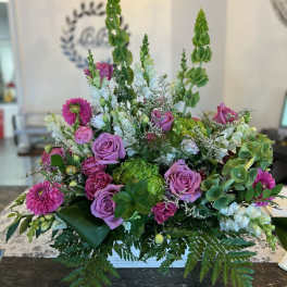 Lush arrangement of purple roses, pink blooms, white flowers, and green Bells of Ireland in a low white container.