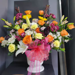 Mixed bouquet of orange roses, lilies, and carnations in a white vase with a pink bow
