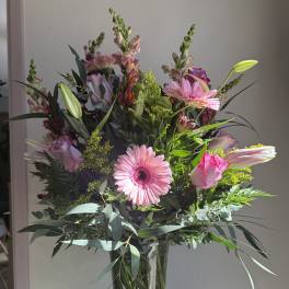 Tall arrangement of pink gerbera daisies, roses, lilies, and snapdragons in a clear glass vase