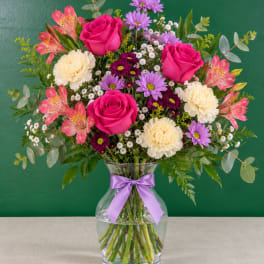 Mixed bouquet of pink roses, carnations, and purple daisies in a glass vase with a ribbon