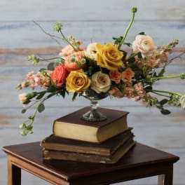 Low arrangement of yellow and peach roses and ranunculus in a silver pedestal bowl on stacked books.