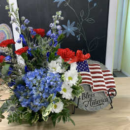 Patriotic floral arrangement with blue hydrangeas, red carnations, and white daisies beside a flag plaque