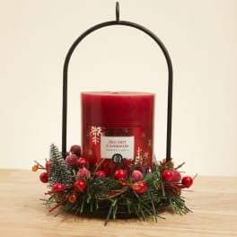 Red scented candle in a black metal handle holder with holiday greenery and berries