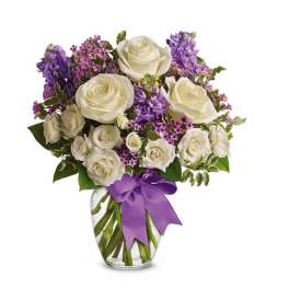 White roses and purple flowers arranged in a clear glass vase with a purple ribbon bow