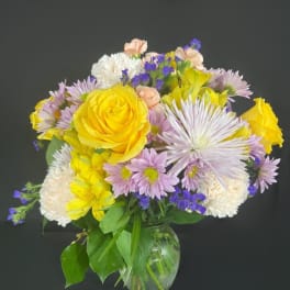 Mixed bouquet of yellow roses, lavender daisies, and white blooms in a clear glass vase
