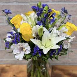 Mixed bouquet of yellow roses, white lilies, and purple flowers in a clear glass vase.