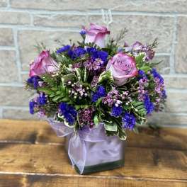 Compact bouquet of pink roses and purple flowers in a square glass vase with a lavender ribbon bow.