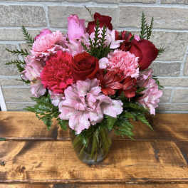 Pink and red roses, carnations, and alstroemeria arranged in a clear glass vase on a wooden table.