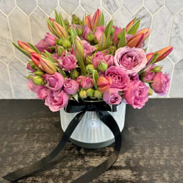 Pink roses and orange tulips arranged in a striped hat box with a black ribbon.