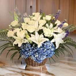 White lilies and roses arranged with blue hydrangeas in a wicker basket
