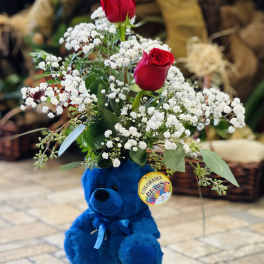 Red roses and baby's breath arranged above a blue teddy bear in a glass vase