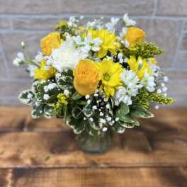 Yellow and white flower arrangement with roses and daisies in a clear glass vase on a wooden table.