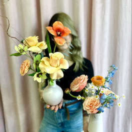 Two small bud vases with peach, yellow, orange, and blue flowers held by a person