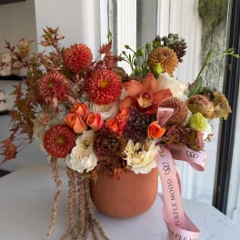 Orange and cream floral arrangement in a terracotta vase with a pink ribbon