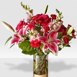 Tall bouquet of pink lilies, red roses, and red carnations in a clear glass vase with gold base.