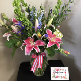 Tall bouquet of pink lilies and blue flowers in a glass vase with a striped ribbon
