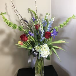 Tall mixed bouquet in a clear glass vase with red roses, blue flowers, and pussy willow stems
