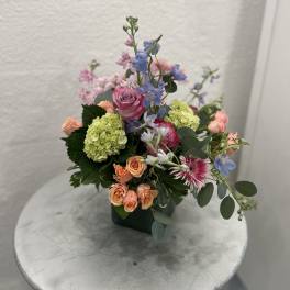 Pastel arrangement of pink and peach roses, green hydrangea and blue blooms in a green square vase