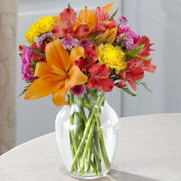 Mixed bouquet of orange, red, pink, and yellow flowers in a clear glass vase